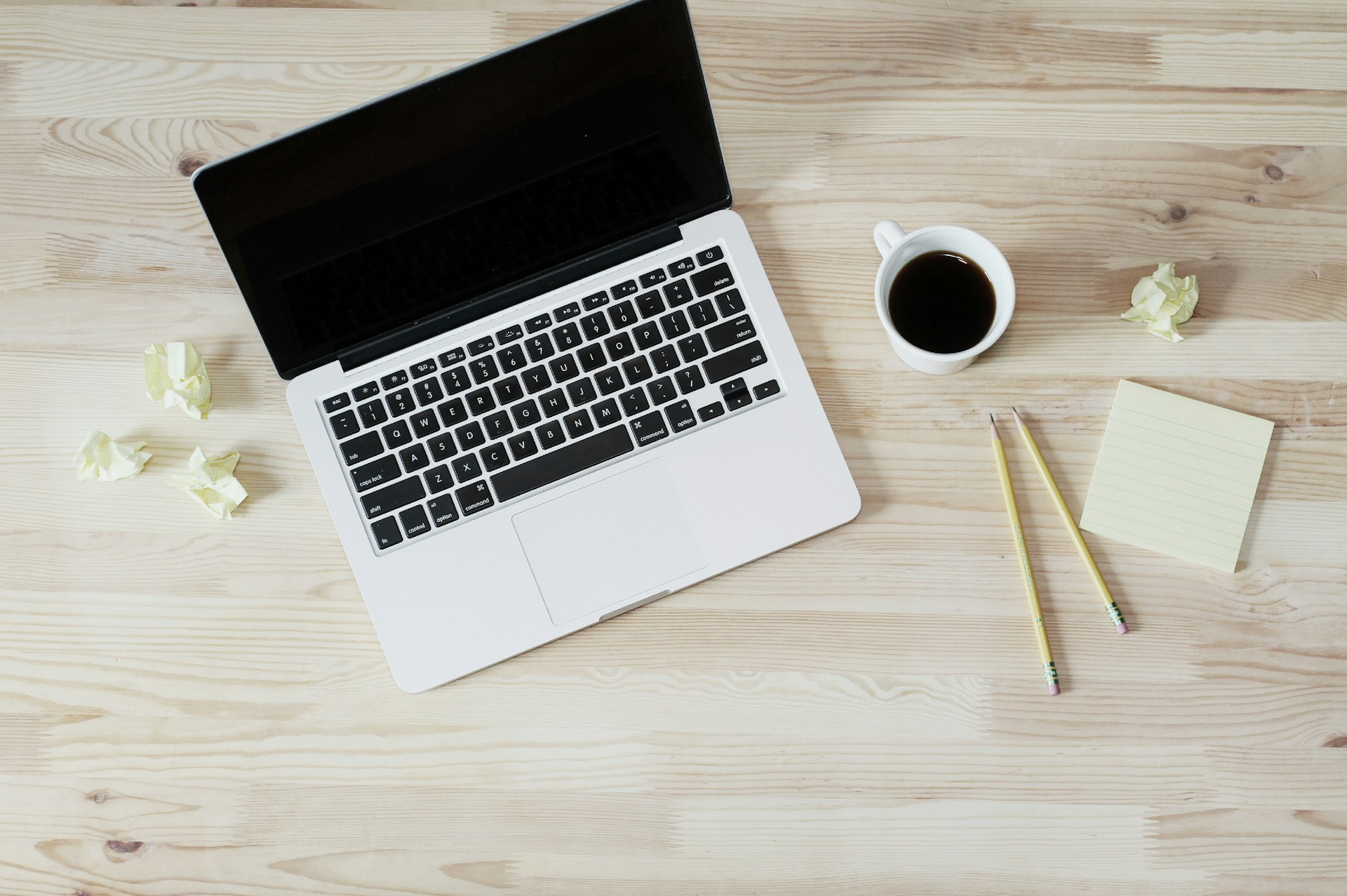 A laptop with a dark screen beside coffee, pencils and note paper on a wooden desk.