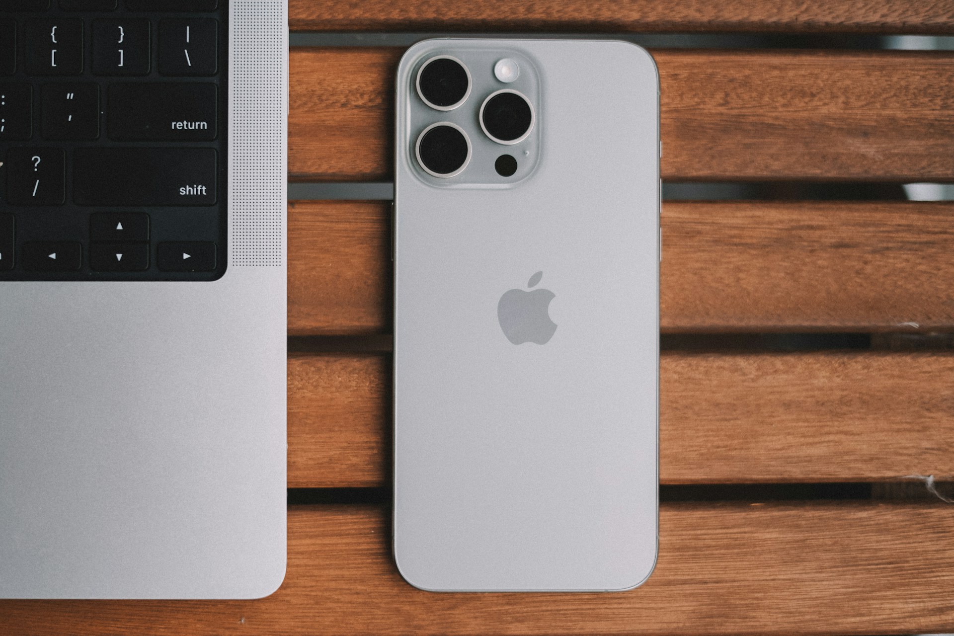 The back of an iPhone on a wooden table beside the edge of a laptop keyboard.